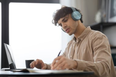 Ciddi bir yönetici, dizüstü bilgisayar kullanıyor ve kulaklık takıyor. Modern ofiste masasında oturuyor. Çevrimiçi Çalışan ve Öğrenen Erkek Serbest Çalışan. Yan Görünüm Görüntüsü, Seçici Odaklanma