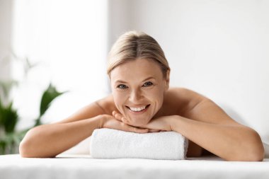 Sağlık Konsepti. Güzel, orta yaşlı bir kadın masaj masasında dinleniyor, olgun bir kadın güzellik tedavisinden hoşlanıyor, lüks bir salonda güzel bir gün geçiriyor, kameraya gülümsüyor, uzay boşluğunu kopyalıyor.