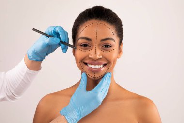 Gülümseyen, güzel, bin yıllık Afro-Amerikan üstsüz kadın estetik ameliyat seansı yapıyor, güzellik uzmanı ya da doktor kadın hasta yüzünü kaplayan tıbbi eldiven giyiyor, gri renkte izole edilmiş.