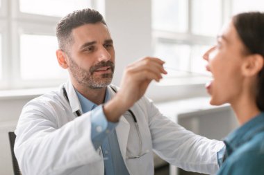 Orta yaşlı erkek doktor, kadının boğazını incelemek için spatula kullanıyor. Terapist, kadının boğazını muayene ediyor.