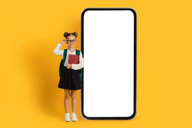 Heyecanlı Küçük Liseli Kız Beyaz Ekranlı Büyük Boş Akıllı Telefonun yanında duruyor, Şirin Gülümseyen Genç Kız Çocuk Eğitim Uygulaması veya Web Sitesi Sarı Arkaplanda Poz Veriyor,
