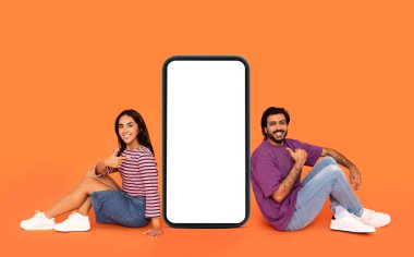 Büyük cep telefonlarıyla yerde oturan, gülümseyen, beyaz ekranı işaret eden, flört uygulaması için model, turuncu stüdyo arka planı, tam boy