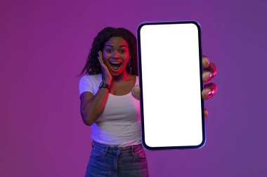 Harika bir teklif. Beyaz Ekranlı Boş Akıllı Telefonu Gösteren Şaşırmış Siyah Kadın, Heyecanlı Afrikalı Amerikalı Kadın Yeni Mobil Uygulama Öneriyor, Mor Arkaplan Üzerinde Neon Işığın Altında Duran, Model