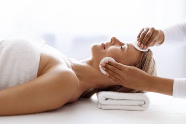 Profesyonel kozmetik uzmanı, güzellik salonundaki cilt bakımı sırasında pamuklu pedlerle orta yaşlı kadının derisini temizliyor. Rahatlamış, olgun, sağlıklı bir kadının Spa Gününün tadını çıkartıyor.