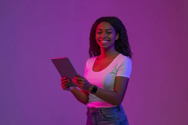 Stock image Digital Leisure. Happy Black Woman With Tablet Computer In Hands Posing In Neon Light Over Purple Background, Smiling African American Female Enjoying Modern Technologies, Copy Space