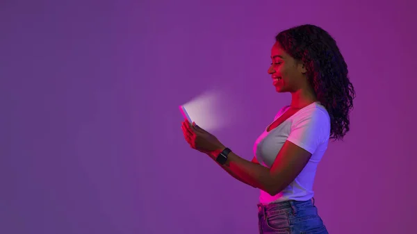 Online Gaming. Smiling Black Woman In Neon Light Looking At Glowing Smartphone Screen, Happy Young African American Lady Holding Mobile Phone, Playing Video Games, Side View With Copy Space