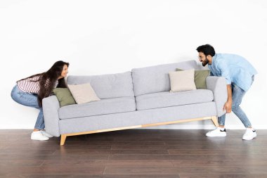 Mutlu, güzel Hintli genç çift, yeni evlerinde beyaz duvar arkası, tam boy, fotokopi alanı üzerinde odada gri kanepeyi taşıyorlar. Taşınma, yeni apartman konseptine geçiş