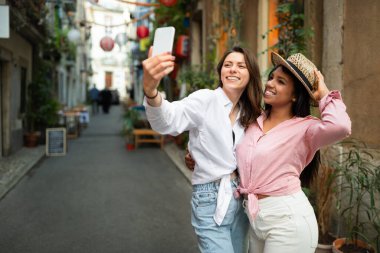 Video görüşmesi, blog. Şapkalı mutlu genç uluslararası kadın turistler telefonda selfie çekiyorlar, sosyal medya için video çekiyorlar ve şehirde maceranın tadını çıkarıyorlar. Tatil ve açık havada seyahat uygulaması