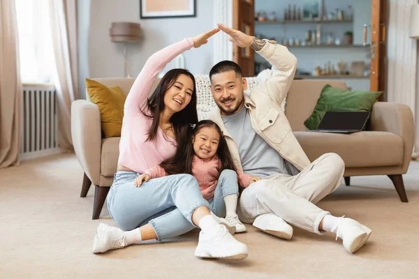 家族住宅 赤ちゃんの娘の上に象徴的な家の屋根を作り 一緒に床に座り 家でカメラに微笑む幸せな日本人の両親 不動産購入と保険 — ストック写真