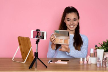 Neşeli, çekici İspanyol kadın günlük kıyafetli güzellik blogcusu canlı yayın yapıyor, profesyonel blog seti ve akıllı telefon kullanıyor, telefon kamerasında kozmetik ürünlerini gösteriyor, pembe arka plan, kopyalama alanı.
