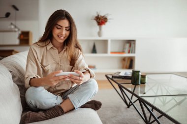 Aygıt, oyun, boş zamanlarında sohbet. Bin yıllık Avrupalı kadın telefonda video izleyip minimalist oturma odasında sörf yapıyor. Çevrimiçi alışveriş, sosyal ağlar, blog ve iletişim uygulaması