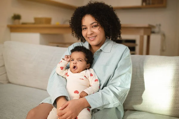 Child care. Portrait of happy pretty young hispanic woman mother with infant baby in arms sitting on couch at home, mom spending time with adorable sleepy curly mixed race toddler child, copy space