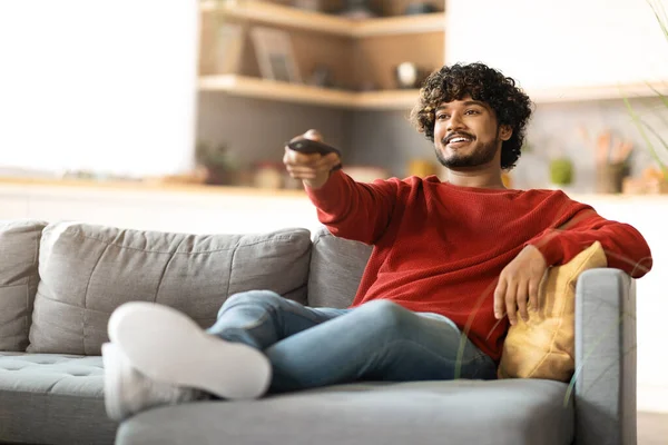 Home Relax. Handsome Indian Guy Sitting On Couch And Switching Tv Channels, Smiling Young Eastern Man Holding Remote Controller, Relaxing On Comfortable Sofa In Living Room, Copy Space