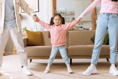 Aile birlikteliği. Anne ve babasıyla el ele tutuşan komik küçük Japon kız, modern oturma odasında ebeveynlerin durduğu kamera pozlarına bakıyor. Kırpılmış Vuruş