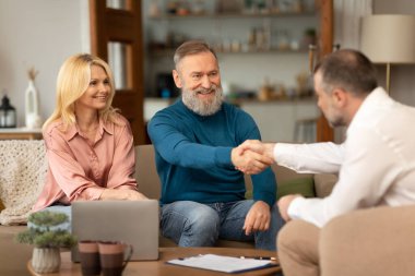 Profesyonel emlakçıyla tokalaşan olgun çift içeride koltukta oturan hizmetlerini sunuyor. Başarılı Anlaşma Konsepti. Neşeli Kıdemli Eşlere Seçici Odaklanma