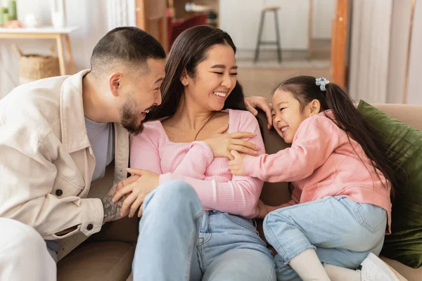 家族の幸せ 陽気な中国人の両親と小さな娘は 自宅でソファに座って遊んでいます ママとパパは週末に彼らの愛らしい子供の女の子のボンディングで遊んで — ストック写真