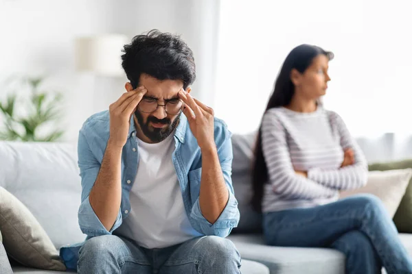 Crisis in relationships. Frustrated stressful young indian man wearing eyeglasses sitting on couch next to girlfriend, touching his head, unhappy guy fighting with wife at home, blurred background