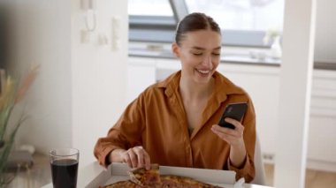 Modern insanların yaşam tarzı. Genç pozitif kadın evde yemek yiyor, pizza yiyor ve sosyal medyada akıllı telefondan internet sörfü yapıyor, çekim takibi, yavaş çekim, serbest alan.