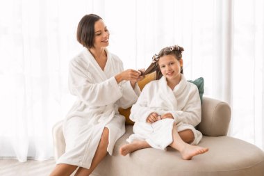 Saç bakım konsepti. Güzel, genç bir anne, küçük kızına bukleler yapıyor, mutlu bir anne ve genç bir kız çocuğu banyodan sonra bornoz giyiyor, birlikte vakit geçiriyorlar.