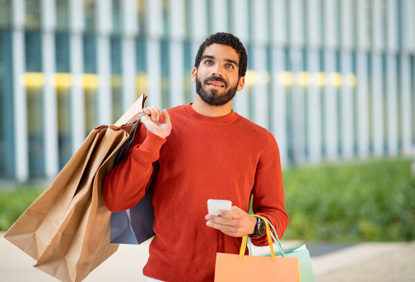 Mobile Shopping Offer. Arabic Guy Using Cellphone Holding Paper Shopper Bags Purchasing New Clothes Online In App Posing Outside. Male Buyer Browsing Internet Via Smartphone Near Mall