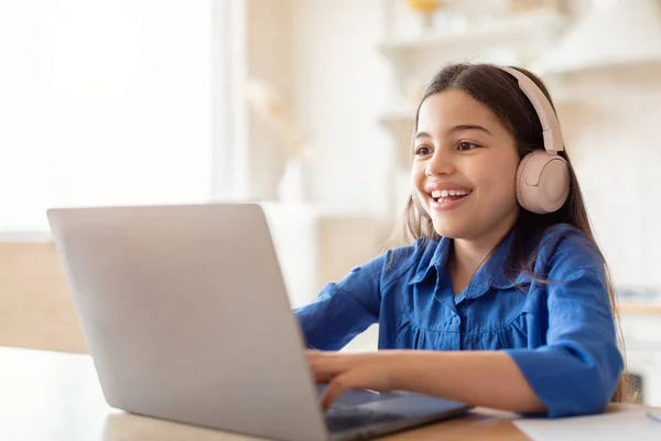 Eラーニング 自宅でヘッドフォンをしてノートパソコンを使ってオンラインで勉強している幸せな女子高生が机に座っています コンピュータをタイプして宿題をする距離クラスを持っている子供の女の子 現代の遠隔教育 — ストック写真