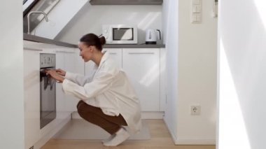 Ev yapımı fırın. Genç pozitif, güzel ev hanımı fırına geliyor ve pastayı kontrol ediyor, sıcaklığı düzeltiyor ve mutfaktan uzaklaşıyor, boş alan.