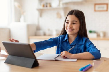 E-öğrenme. Dijital tablet kullanan, internette ödev yapan ve evde masada oturan mutlu küçük liseli kız. Arapça çalışan kız bilgisayarın yanında not alıyor. Teknoloji ve Eğitim
