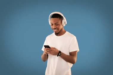 Havalı, mutlu, gülümseyen Afrikalı Amerikalı genç adam yeni kablosuz kulaklıklar ve akıllı telefonlar kullanarak mavi stüdyo arka planında video içeriğini internetten izliyor, kopyalama alanı izliyor. Modern teknolojiler