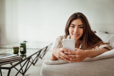 İş için uygulama ve cihaz. Aydınlık oturma odasının içinde telefonla yazan Avrupalı olumlu bir kadın. Evde sosyal medya sohbeti, iyi mesaj, haber, oyun ve arama