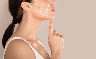 Cropped of young woman with clean fresh skin touching her chin, antiaging concept. Side view of unrecognizable lady in white top with lifting arrows on cheek, beige studio background, copy space clipart