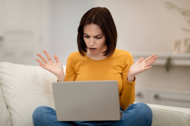 Duygusal olarak sinirli genç esmer kadın evdeki kanepede oturuyor, dizüstü bilgisayara bakıyor ve jestler yapıyor, dijital göçebe bilgisayar yazılımlarıyla ilgili sorunlar yaşıyor, kopyalama alanı