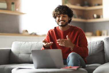 Gülümseyen Hintli genç adam evde dizüstü bilgisayarla video görüşmesi yapıyor, yakışıklı doğu çocuğu oturma odasında oturuyor ve online iletişim için bilgisayar kullanıyor, web kamerasında konuşuyor, uzayı kopyalıyor.