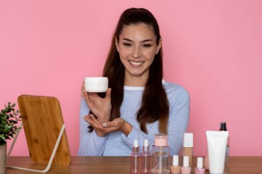 Güzel, uzun saçlı, esmer bir kadın günlük kıyafetlerle kozmetik uzmanı masasında çeşitli güzellik ürünleri ve aynalarla oturuyor. Yüz kremi ve pembe arka planda beyaz kavanozu gösteriyor.