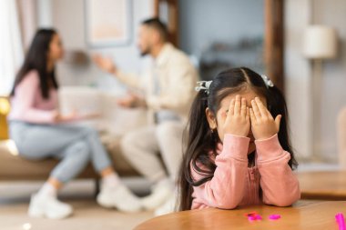 Aile çatışması. Küçük Asyalı bir kız, ebeveynler evdeki koltukta otururken birbirlerine bağırırken gözlerini kapayarak ağlıyor. Depresyondaki bebeğe odaklan. Mutsuz Çocukluk Konsepti