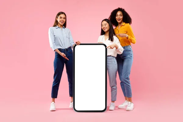 酷在线报价 快乐的三位女士指着粉色背景上的空白大智能手机屏幕 模仿着 快乐女性推荐新的移动应用程序或网站 — 图库照片