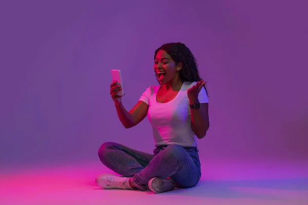 Good News. Excited Black Woman Looking At Smartphone Screen And Shouting Yes, Cheerful Young African American Female With Mobile Phone Sitting On Floor In Neon Light On Purple Background