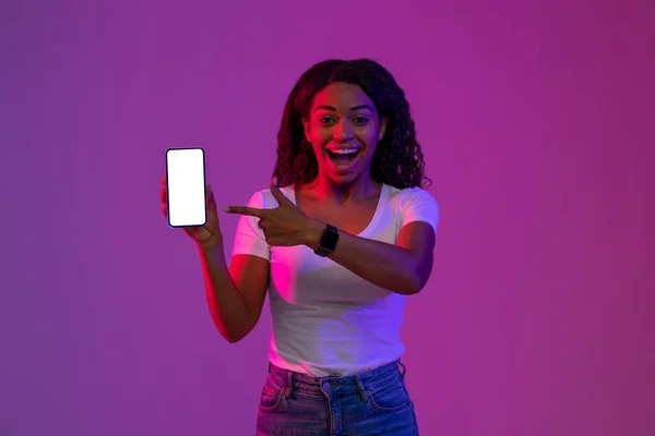 移动广告 兴奋的黑人女人拿着白屏的空白智能手机指指指点点 站在紫色背景的霓虹灯下推荐新的应用程序或网站 — 图库照片
