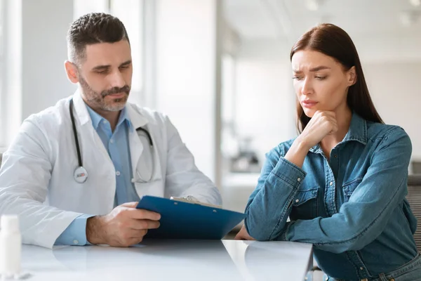 Male physician discussing check up results with worried woman during meeting at hospital, female patient touching chin while doctor explaining her details of treatment