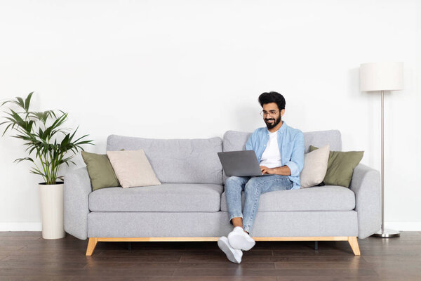 Freelance. Cheerful millennial indian man in casual outfit wearing eyeglasses sitting on sofa over white wall, typing on laptop keyboard and smiling, guy digital nomad working from home, copy space