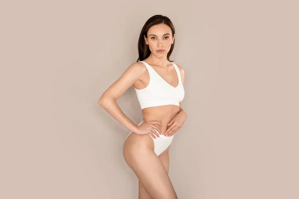 Confident beautiful brunette millennial woman posing in white comfortable underwear on beige studio background, showing perfect, well-fit young body, copy space. Healthy lifestyle for women