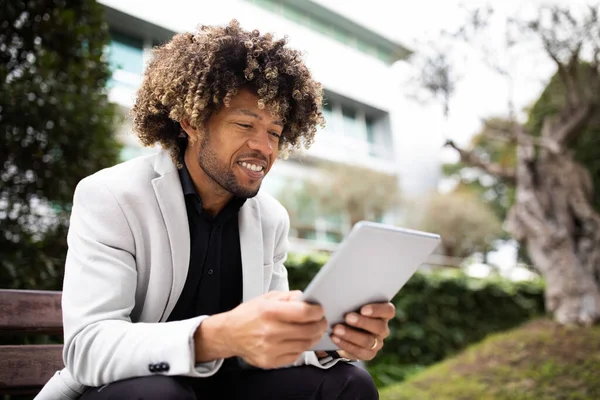 Happy confident black middle aged businessman in suit using digital tablet, sitting on bench near office. Successful business outdoor, man using app for trade, ad and offer