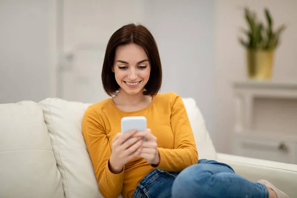 很好的提议穿着休闲装的快乐漂亮的黑发女青年独自坐在沙发上 使用全新的智能手机 查看社交媒体 与朋友聊天 复制空间 — 图库照片