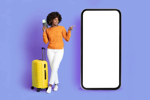 兴高采烈的年轻黑人女性展示移动应用程序或网上提供旅游产品 非洲裔女士带着行李 指向装有白屏的大型智能手机的票 紫色背景 — 图库照片