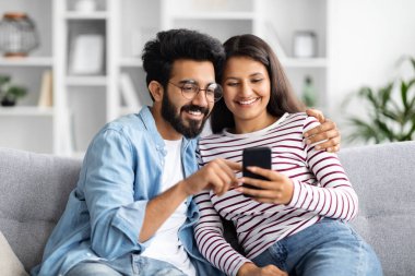 Relaxed cheerful beautiful young multicultural couple middle eastern guy and indian woman sitting on sofa at home, using smartphone and smiling, shopping online, surfing on Internet, copy space clipart
