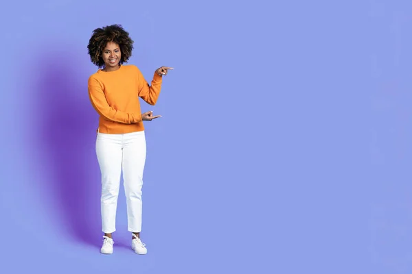 いい取引だ 陽気スタイリッシュな美しいです若いアフリカ系アメリカ人女性で素敵な衣装を指して脇 上の広告のためのコピースペースを示します紫色のスタジオの背景 フル長さのショット — ストック写真