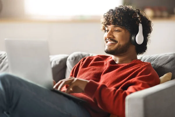 Smiling Indian Freelancer Guy Working On Laptop Online At Home And Listening Music In Wireless Headphones, Handsome Eastern Man Sitting On Couch With Computer, Enjoying Remote Job, Copy Space