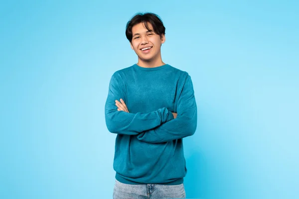 Confident Japanese Teen Guy Posing Crossing Hands And Smiling To Camera Over Blue Studio Background. Shot Of Cheerful Teenager Student Standing Wearing Casual Clothes. Self Confidence