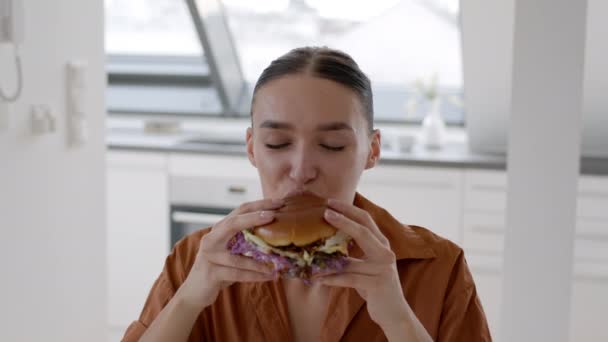 垃圾食品爱好者 近照年轻快乐女人的画像 她咬着新鲜美味的大汉堡 舔着手指 在家里吃着骗人的饭菜 对着相机微笑 慢动作 — 图库视频影像