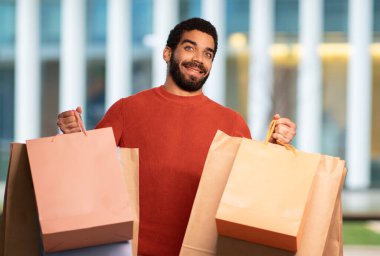 Modern AVM 'nin dışında duran fotoğraf makinesine kağıt torbaları gösteren heyecanlı bir Arap adam. Erkek Alışverişi Holding Paketleri Reklam Satışı Teklifi. Ticaret ve Perakende Konsepti