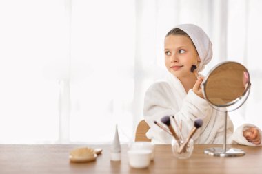 Evde giyinme masasında otururken makyaj yapan sevimli küçük kız, bornoz takan, utangaç fırça kullanan ve fotokopi odasına yan bakan mutlu kız çocuğu,